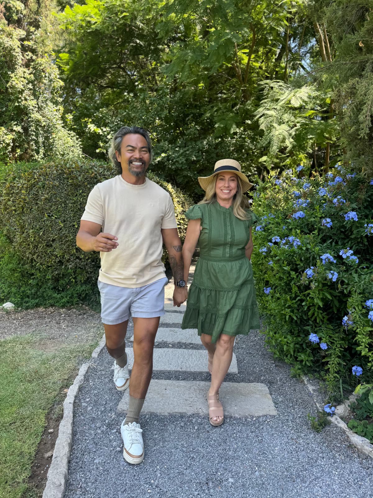 Tommy and Lety walking hand in hand down a garden path lined with greenery and blue flowers.