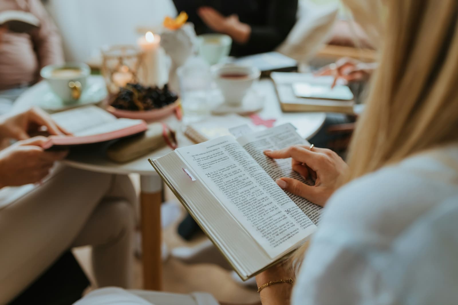 A woman reading an open Bible at a table during a small-group study, coffee and pastries in front of her.