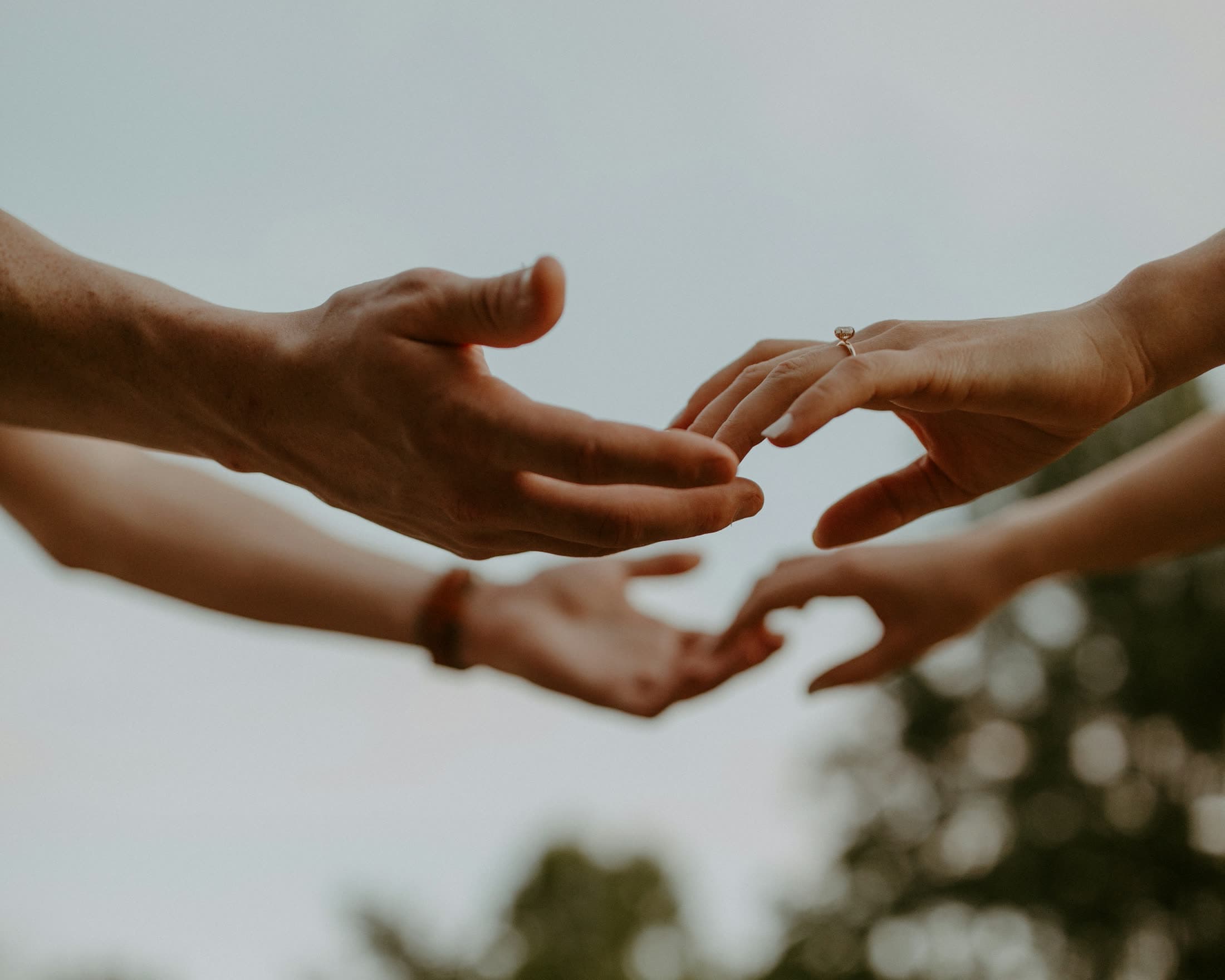 Two pairs of hands reaching toward one another against a pale sky, one wearing an engagement ring.