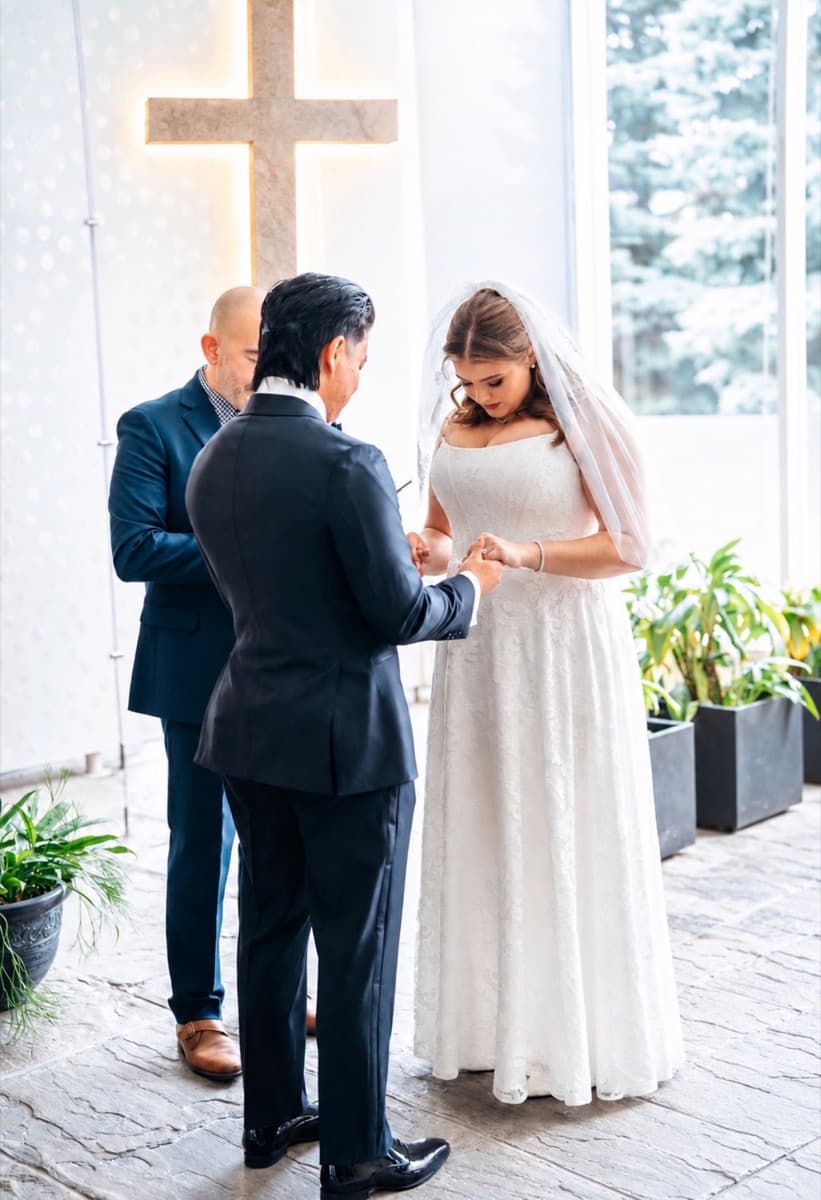 A bride and groom exchanging rings before an officiant, a lit cross on the wall behind them.